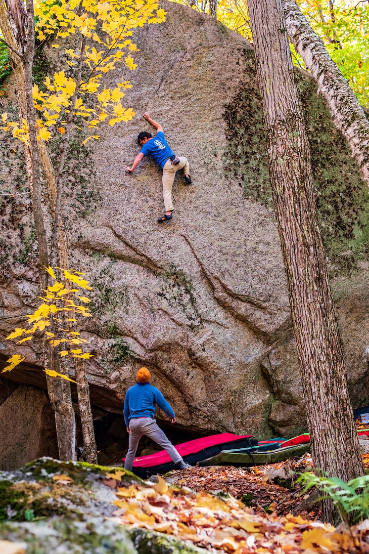 Climbing on a highball boulder problem while spotter adjusts pads