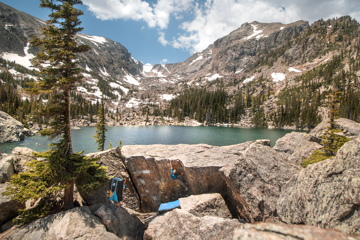 tanner mad rock 1 White man in blue shirt climbs the side of a boulder with Lake Haiyaha in the background.