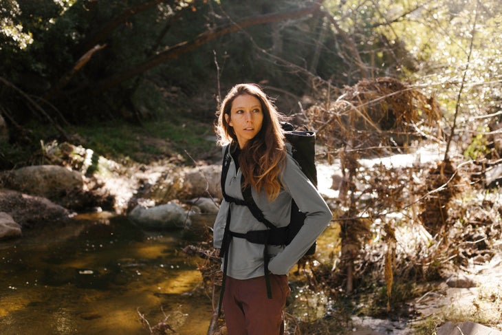 BCAndyB_190222_0060 White, brunette woman looks off camera while wearing a gray hoodie and maroon climbing pants. A black backpack sits on her back. There is a creek and…