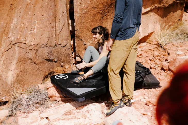 BCAndyB_190226_0109 Man in blue shirt and beige pants stands next to a crash pad where a brunette white woman sits and ties her climbing shoes.