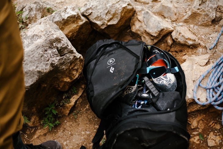 Bokanev-BGAClimbMex-4291 Black Backcountry and Black Diamond backpack sits on ruddy rock unzipped showing climbing shoes and a belay device.