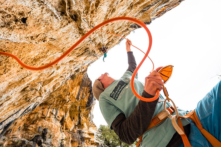 best-belayer_grigri White man in blue clothes feeds slack to a climber. Photo shot from below looking up at the belayer and climber.