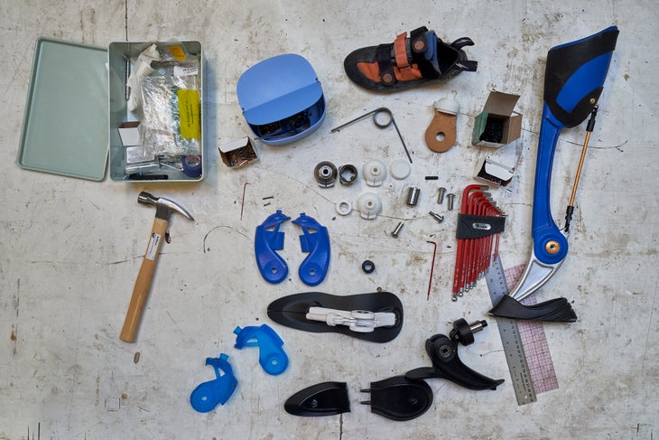 190301_KaiLin_NYC_Percival_09410 Parts to create a prosthetic leg lie on a dirty white background: a hammer, tin box with plastic wrappers, a climbing shoe, screws, plastic parts,…