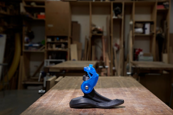 190301_KaiLin_NYC_Percival_09963 Prosthetic climbing foot sits on a wooden table in a workshop. Bottom of the foot is black and curved like a foot. The ankle joint is blue with a…