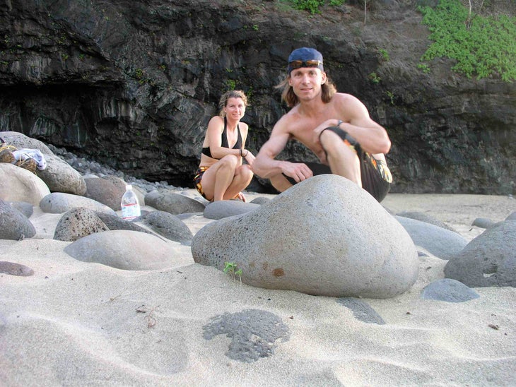 Chalk Cartel owners and climbing chalk experts Timy and Brandi pose crouched on a beach in Hawaii, a dark overhung cliff behind them.