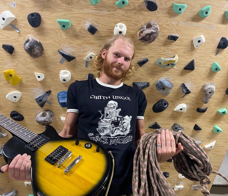 holding guitar and climbing rope in hands. In front of a climbing wall.