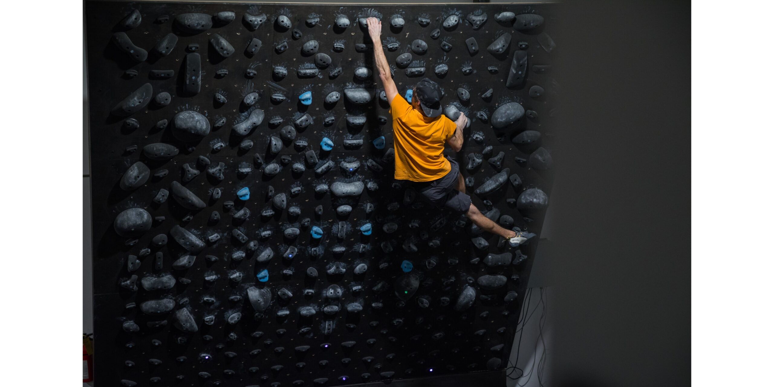 Man in orange shirt climbs on a black bouldering wall with lightened holds.