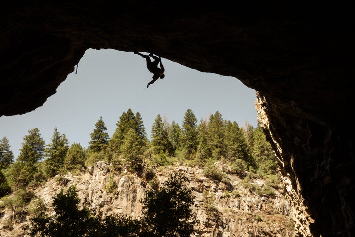 Siegrist in silhouette hanging from the lip of the cave, pine trees and growing from the top of the canyon's opposite rim.