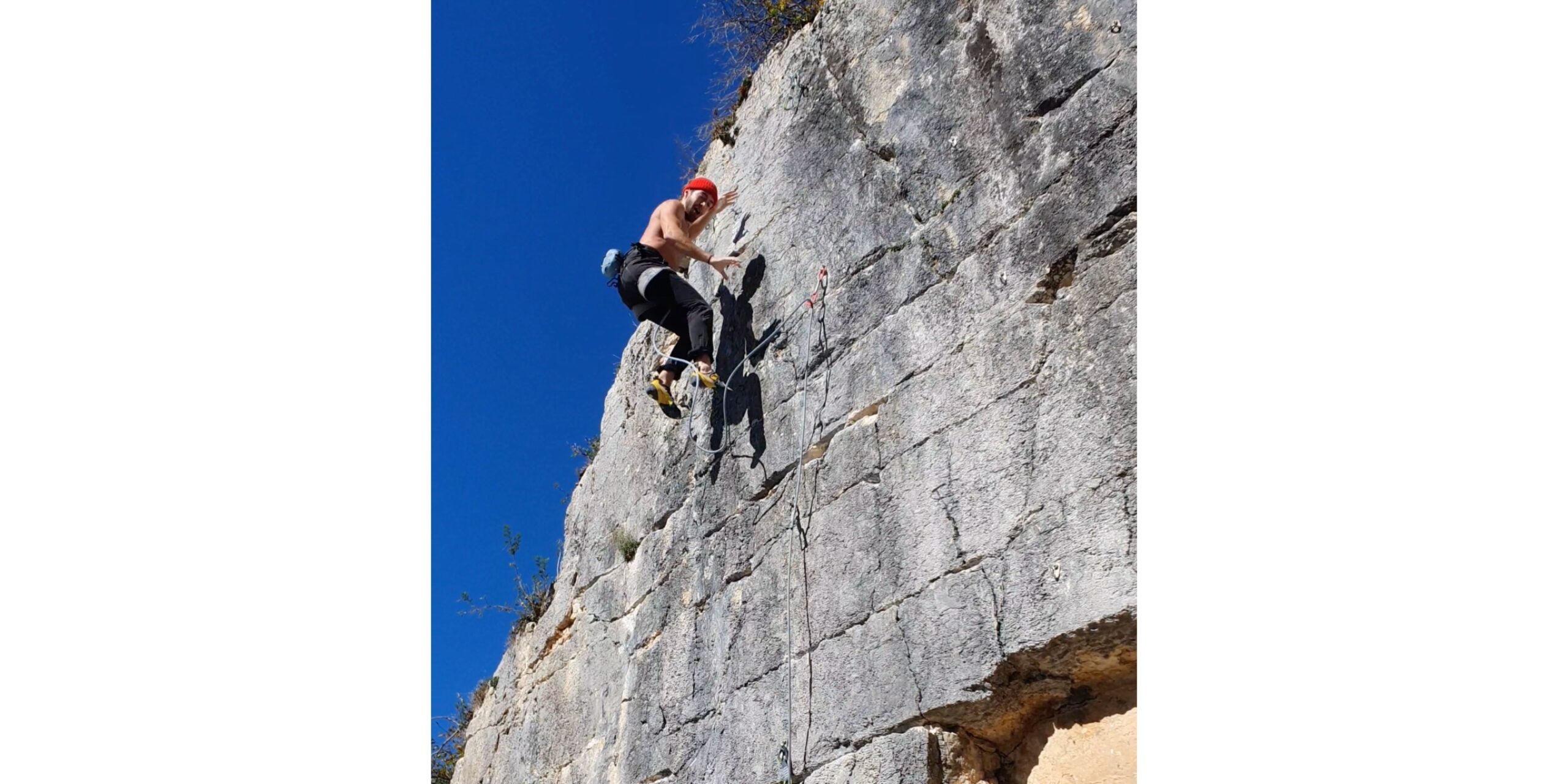 Rock climber falls down vertical rock cliff with rope wrapped around right leg. Sunny, blue-sky day.